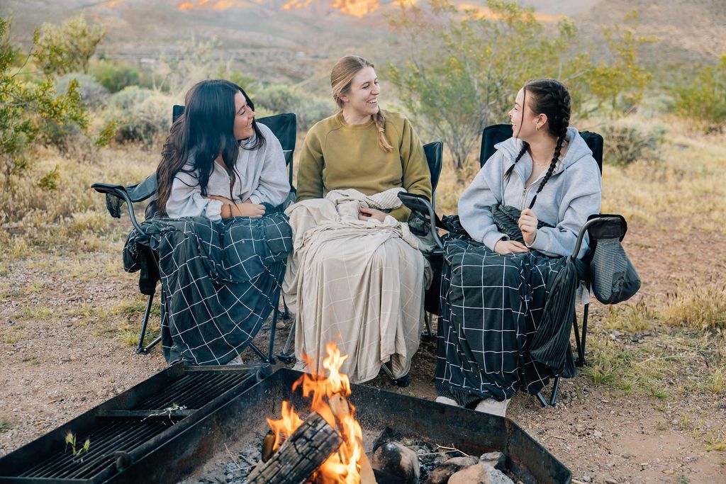 Three people sitting on camping chairs by a fire with charcoal and light tan grid pattern blankets on each of them.