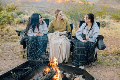 Three people sitting on camping chairs by a fire with charcoal and light tan grid pattern blankets on each of them.