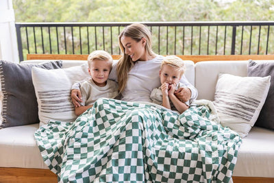 A mother and two children sitting on a couch with a white and green checkered printed blanket over all three of them.