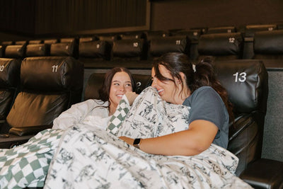 Two people in movie theater seats with a green and white checkered blanket and a western print blanket on each of them. 