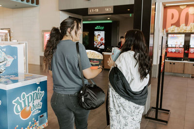 Two people in a movie theater holding a popcorn bucket and drink as well as a black rounded bag and a western printed blanket.