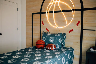 A sports themed bdroom with a twin bed made with blue sports themed kids zipper bedding and a wall decorated to look like the floor of a basketball court