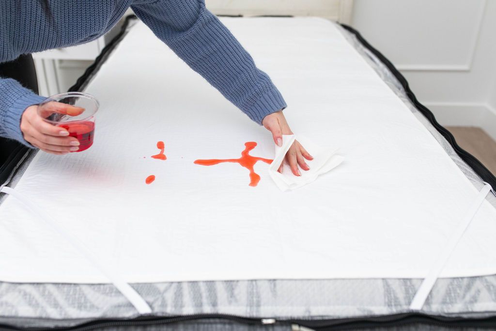 White absorbent accident bedding pad with red liquid poured on top and someone wiping it off the bed with a paper towel