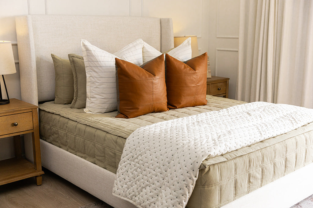A queen bed in a brightly lit bedroom made with khaki green zipper bedding and an assortment of cream and leather bedding accessories.