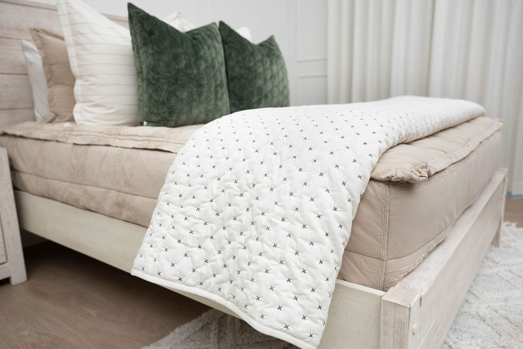 A side view of a bed made with tan zipper bedding with a cream embroidered blanket folded over the end of the bed.