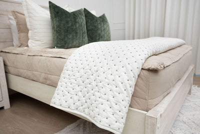 A side view of a bed made with tan zipper bedding with a cream embroidered blanket folded over the end of the bed.