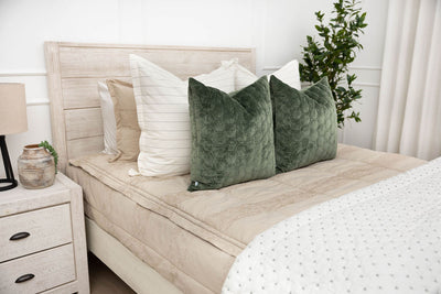 A brightly lit bedroom with a bed made with tan zipper bedding and neutral and velvet green pillows and blanket.