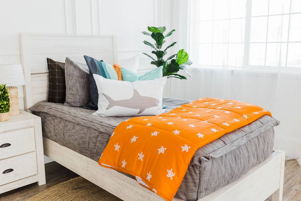 Brown and gray woven textured bedding, white, teal, orange, white striped euro, teal pillow with white shark print, white lumbar with gray shark print, and orange blanket with white turtle print at the foot of the bed