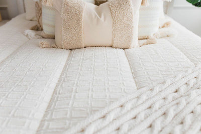 Cream bedding with textured rectangle design and dark creamy textured euro, a cream and tan woven textured pillow and a textured dark creamy lumbar with tassels with an off white braided throw with pom poms along the edge