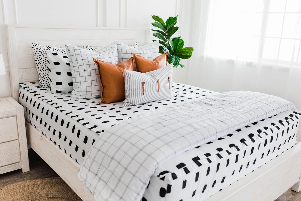 Queen bed with White bedding with black dashed lines and white and black grid euro, faux leather pillows, white and black striped lumbar with faux lather buckles and white and black grid pattered blanket