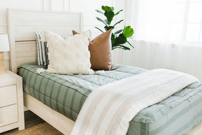 Twin bed with green striped bedding, faux leather euro, cream euro with textured polka dots and tassels on the corners, with a cream blanket with woven brown stripes