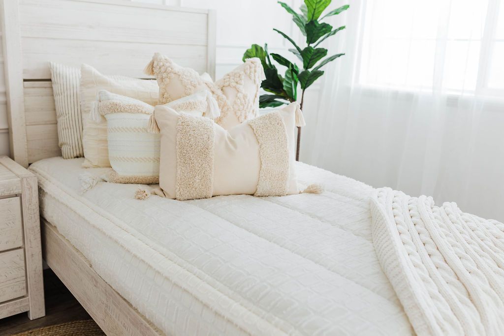 twin bed with Cream bedding with textured rectangle design and dark creamy textured euro, a cream and tan woven textured pillow and a textured dark creamy lumbar with tassels with an off white braided throw with pom poms along the edge