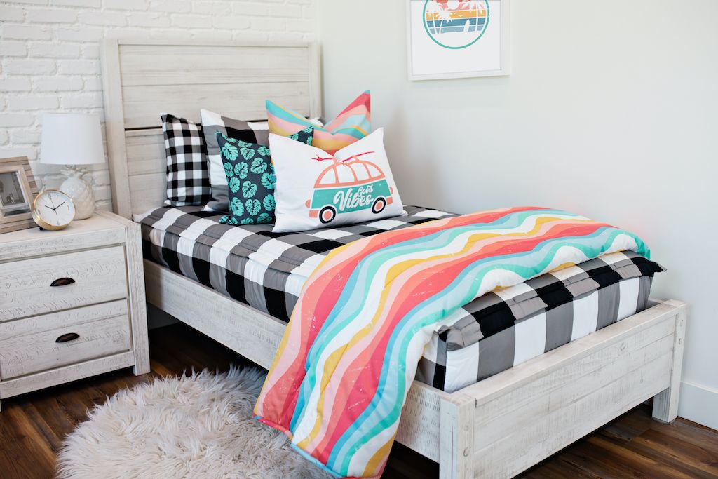 twin bed with Black and white buffalo plaid bedding and pink, blue, yellow toned wavy lined euro, blue pillow with teal palm leaves, and white lumbar coral and teal beach bus, blue, yellow wavy lined blanket at the foot of the bed