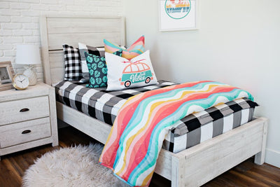twin bed with Black and white buffalo plaid bedding and pink, blue, yellow toned wavy lined euro, blue pillow with teal palm leaves, and white lumbar coral and teal beach bus, blue, yellow wavy lined blanket at the foot of the bed