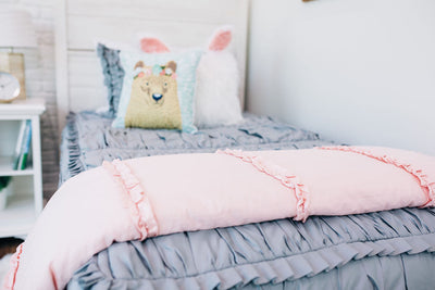 White twin size bed with gray ruffled bedding and a blush pink ruffled blanket at the foot of the bed.