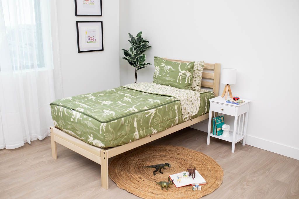A twin bed made with olive green dinosaur fossils kids zipper bedding, a nightstand beside it with books on the shelf and toys scattered on the floor.