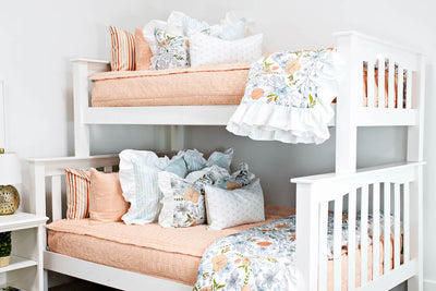 bunk bed with Peach bedding with textured rectangle design with white and blue striped euro with ruffle along the edge, floral printed pillow, white lumbar with small gray floral design and floral printed blanket with white ruffle along the edge