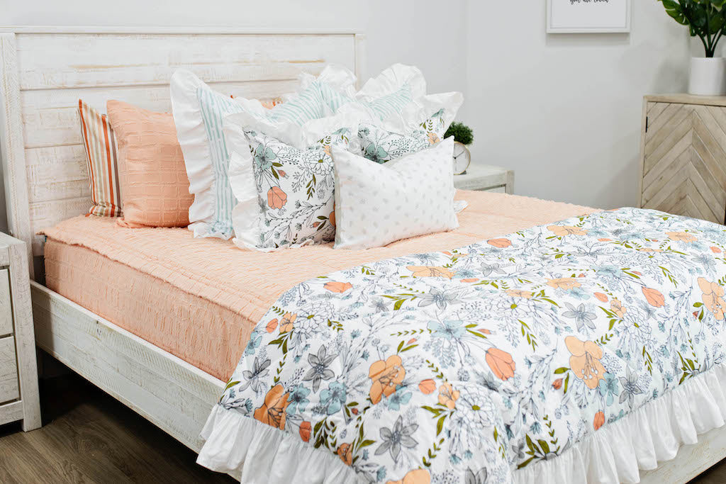 queen bed with Peach bedding with textured rectangle design with white and blue striped euro with ruffle along the edge, floral printed pillow, white lumbar with small gray floral design and floral printed blanket with white ruffle along the edge