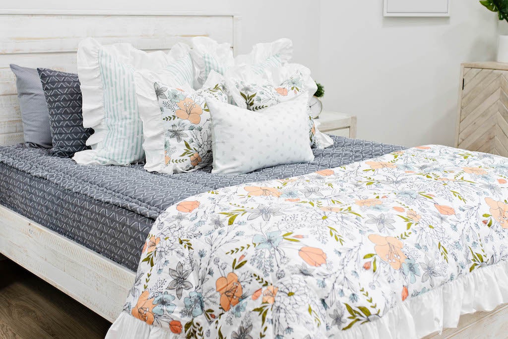 queen bed with Gray bedding with textured diamond white and blue striped euro with ruffle along the edge, floral printed pillow, white lumbar with small gray floral design and floral printed blanket with white ruffle along the edge