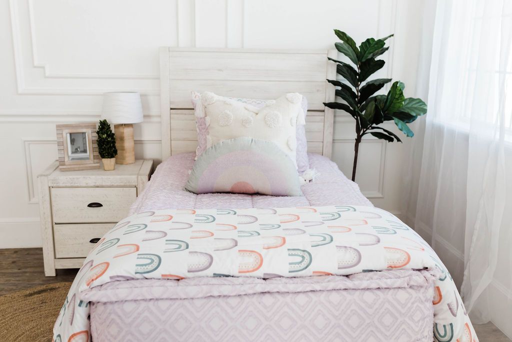 White bedframe, textured purple zipper bedding with textured euro pillow with tassels, pastel rainbow pillow, and a white blanket with ombre purple, orange and green rainbows