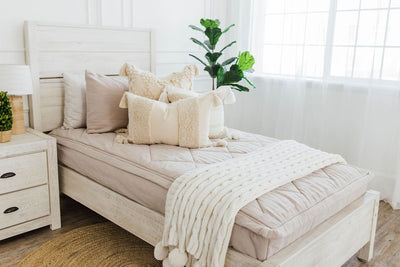 twin bed with Tan textured zipper bedding dark creamy textured euro, a cream and tan woven textured pillow and a textured dark creamy lumbar with tassels with an off white braided throw with pom poms along the edge