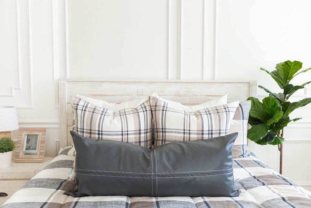 A brightly lit bedroom with a bed made with cream, tan, and charcoal plaid zipper bedding and an assortment of neutral and leather pillows.
