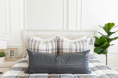 A brightly lit bedroom with a bed made with cream, tan, and charcoal plaid zipper bedding and an assortment of neutral and leather pillows.