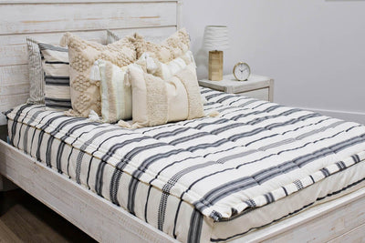 queen bed with  cream and black woven striped bedding with dark creamy textured euro, a cream and tan woven textured pillow and a textured dark creamy lumbar with tassels