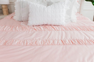 Blush pink bedding with white ruffle polka dot euros,  ruffle gray/blue textured pillows, white ruffle textured lumbar, and a textured white throw with braided tassels