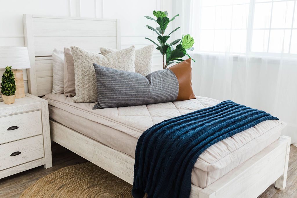 twin bed with Tan textured zipper bedding, dusty tan faux fur euro, blue striped and faux lather XL lumbar with navy knitted chenille throw with pom poms