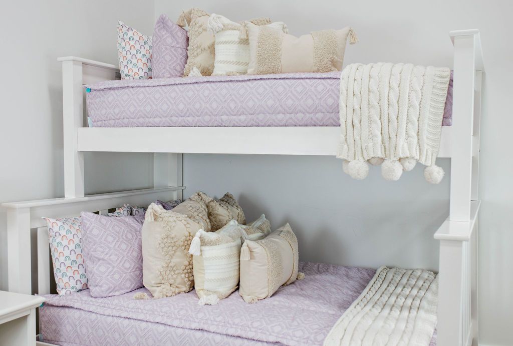 White bunk bed with purple textured bedding, dark creamy textured euros, cream and tan woven textured pillows and a textured dark creamy lumbar with tassels with an off white braided throw with pom poms along the edge