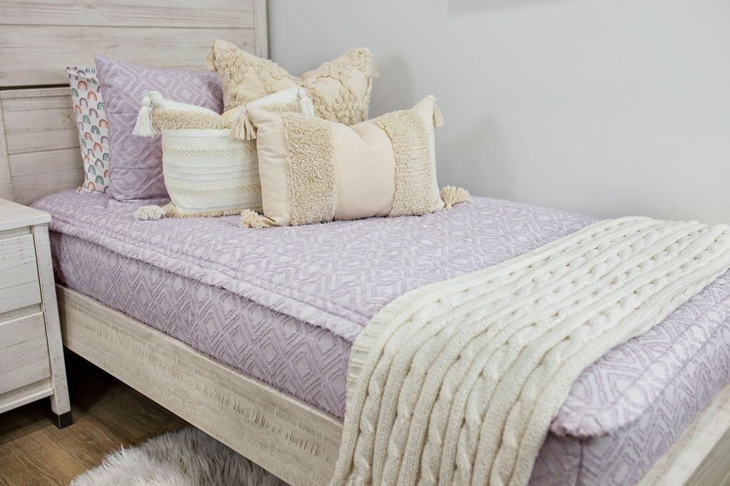 White twin bedframe with a dark creamy textured euro, a cream and tan woven textured pillow and a textured dark creamy lumbar with tassels with an off white braided throw with pom poms along the edge