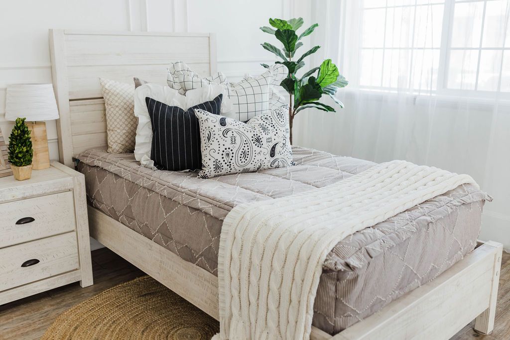 twin bed with Taupe bedding with textured zig zag design and cream and black striped euros with ruffle along the edge, charcoal striped pillow with white ruffle along the edge, cream lumbar with charcoal paisley print, and cream textured blanket with pom poms