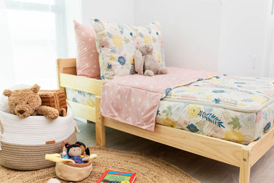 A toddler bed made with pink, yellow, and blue floral kids zipper bedding and a basket of stuffed animals and toys beside it.