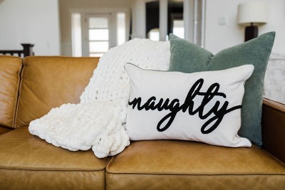 White pillow with black text spelling 'naughty' with green pillow and white throw blanket