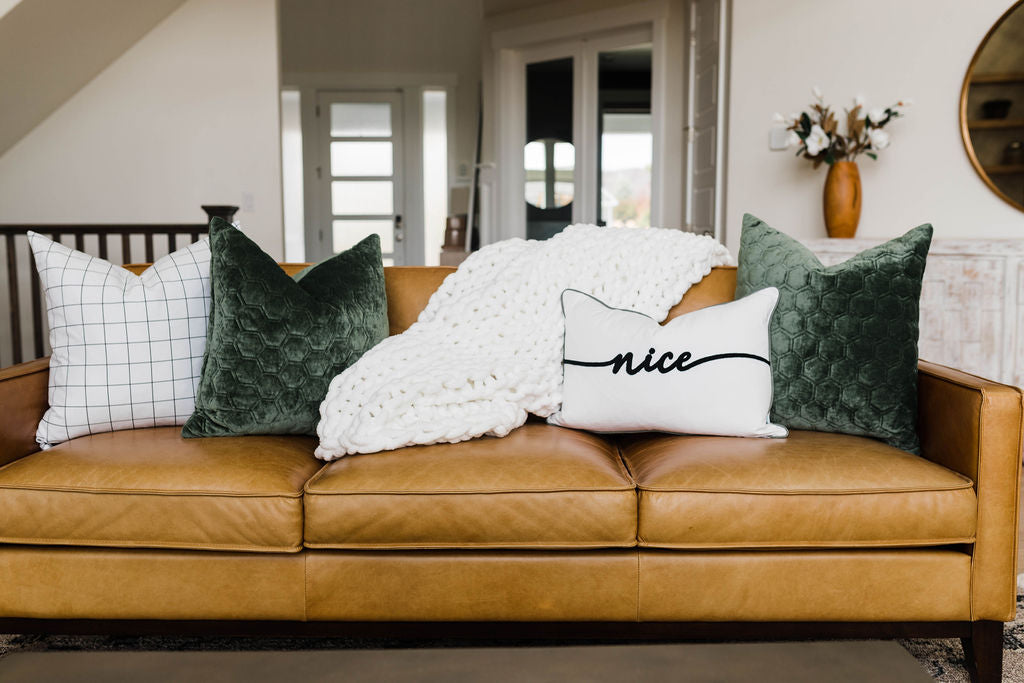 White pillow with black text spelling 'nice' on tan couch with green and white pillows and a white throw blanket