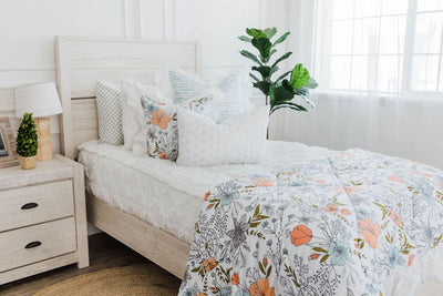White bedding with texturized diamond design with , white and blue striped euro with ruffle along the edge, floral printed pillow, white lumbar with small gray floral design and floral printed blanket with white ruffle along the edge