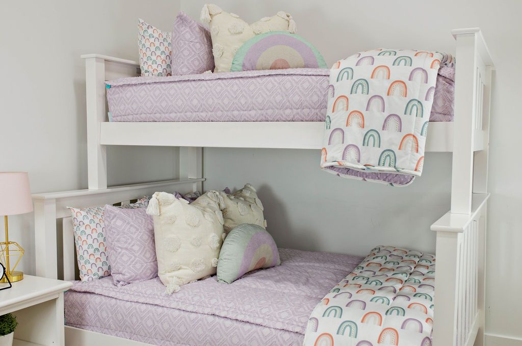 White bunk bed with purple textured bedding and textured euro pillow with tassels, pastel rainbow pillow, and a white blanket with ombre purple, orange and green rainbows
