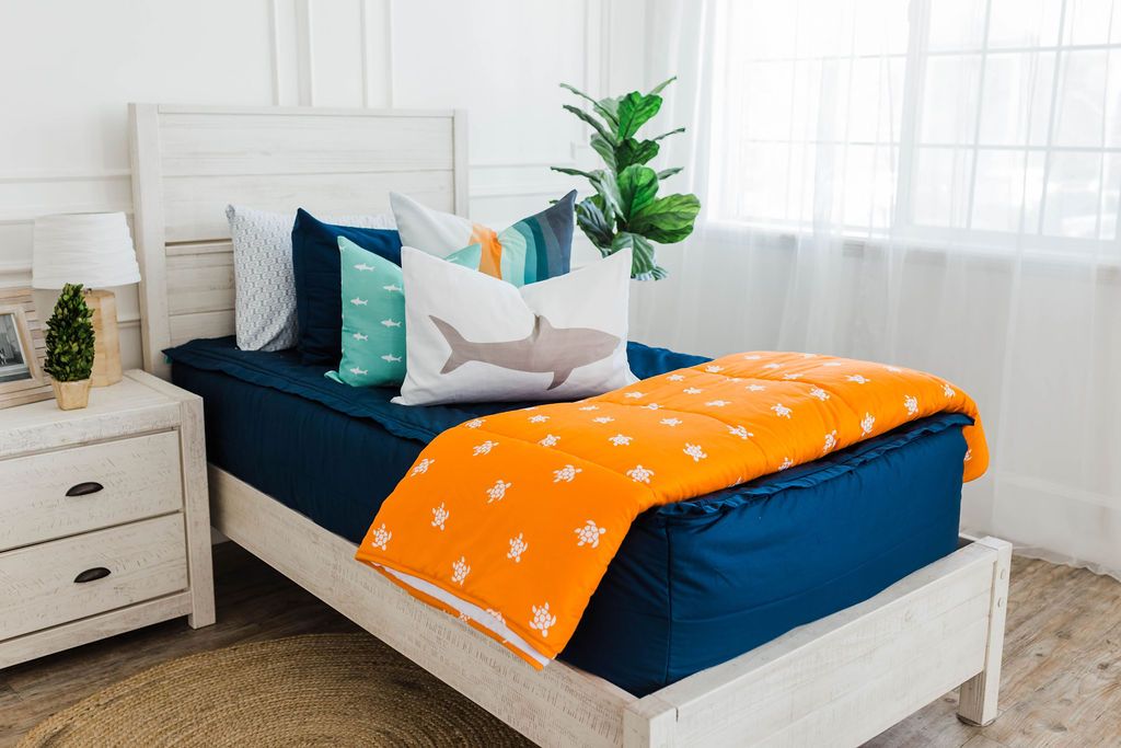 twin bed with Navy blue zipper bedding with white, teal, orange, white striped euro, teal pillow with white shark print, white lumbar with gray shark print, and orange blanket with white turtle print at the foot of the bed