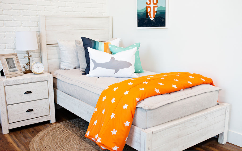 twin bed with tan textured bedding and white, teal, orange, white striped euro, teal pillow with white shark print, white lumbar with gray shark print, and orange blanket with white turtle print at the foot of the bed