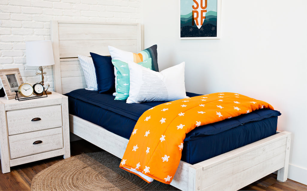 twin bed with navy zipper bedding and white, teal, orange, white striped euro, teal pillow with white shark print, white lumbar with gray shark print, and orange blanket with white turtle print at the foot of the bed