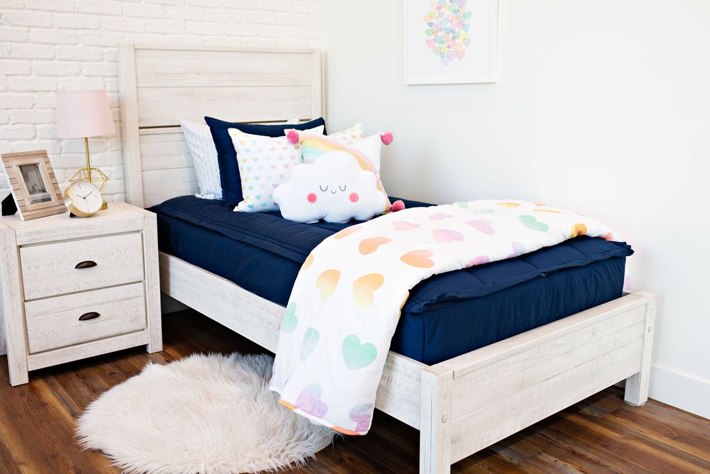 White twin size bed with navy blue bedding, a medium rainbow heart pillow, a medium rainbow pillow, a white cloud shaped pillow and a white and rainbow heart blanket.  