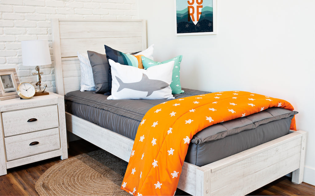 twin bed with gray zipper bedding and white, teal, orange, white striped euro, teal pillow with white shark print, white lumbar with gray shark print, and orange blanket with white turtle print at the foot of the bed