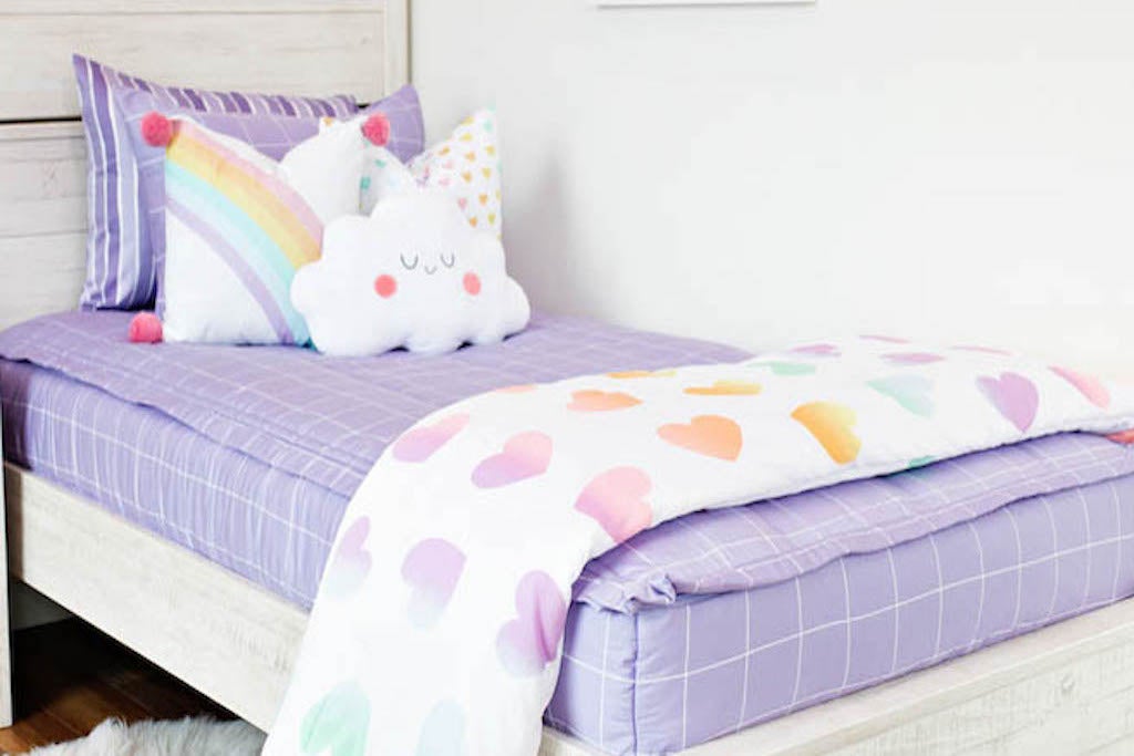 twin bed with Purple bedding with white grid pattern, white pillow with rainbow ombre hearts, white pillow with rainbow and pink pom poms on the corner, plush marshmallow pillow, and white blanket with ombre hearts at the foot of the bed