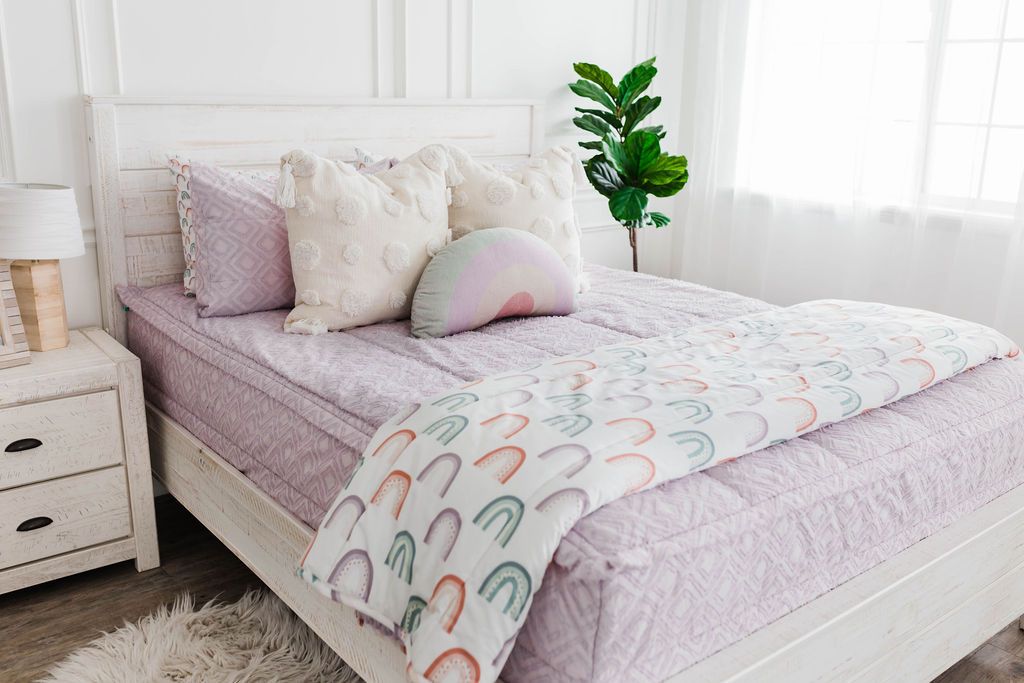 White queen bedframe, purple textured zipper bedding and textured euro pillow with tassels, pastel rainbow pillow, and a white blanket with ombre purple, orange and green rainbows