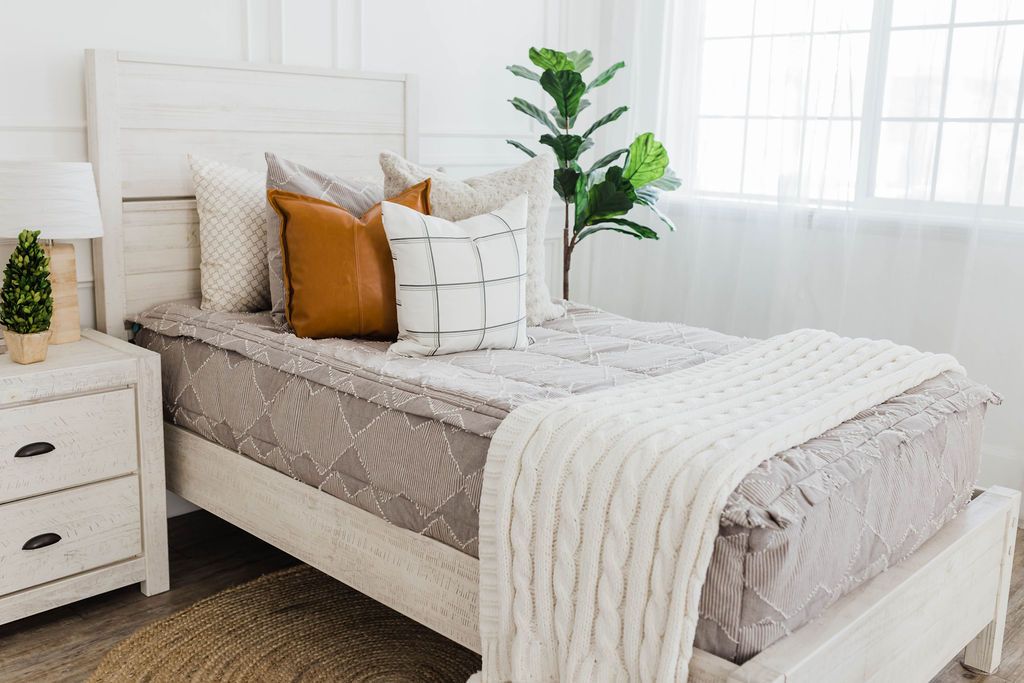 twin bed with Taupe bedding with textured zig zag design and dusty tan faux fur euro, faux leather pillow, white pillow with dark gray grid pattern, and cream knitted chenille throw with pom poms