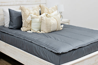 queen bed with Gray zipper bedding with dark creamy textured euros, cream and tan woven textured pillows and a textured dark creamy lumbar with tassels 