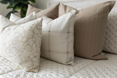 A close up side view of a bed made with cream embroidered zippered bedding and an assortment of pattern neutral pillows.