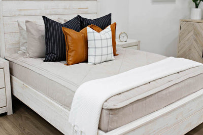 queen bed with Tan textured zipper bedding deep navy striped euros, faux leather pillows, white and black grid pillow, and white textured blanket with braided tassels