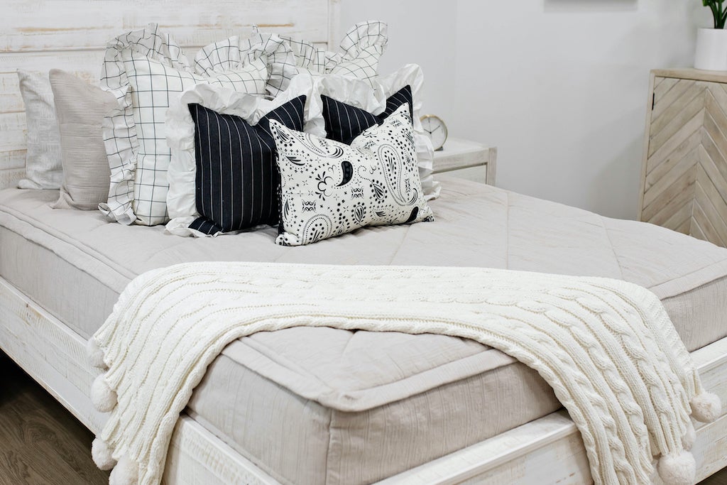 queen bed with Tan textured zipper bedding, cream and black striped euros with ruffle along the edge, charcoal striped pillow with white ruffle along the edge, cream lumbar with charcoal paisley print, and cream textured blanket with pom poms